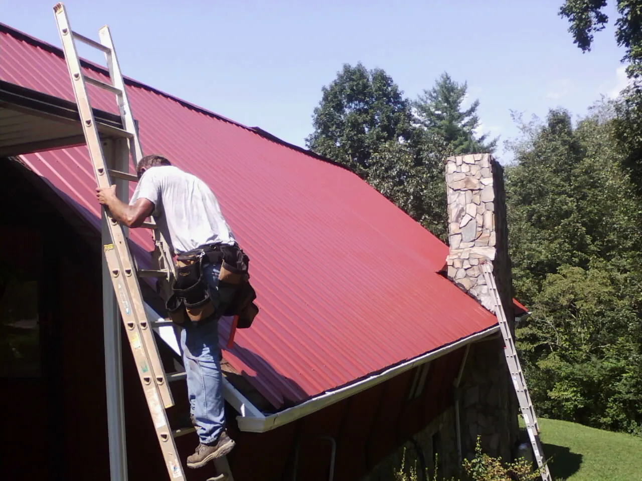 Roofer on a ladder beside a red metal roof and stone chimney — Soffit & Fascia Repair by Roofing Brothers in Sour Lake